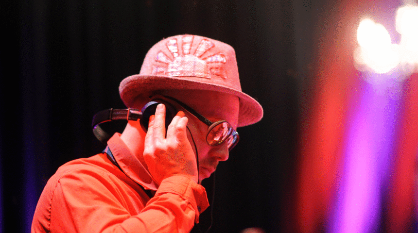 Yuriy Gurzhy mit rotem Hemd und rotem Hut steht an einem DJ-Pult vor einem schwarzen Vorhang mit Kronleuchtern im Hintergrund